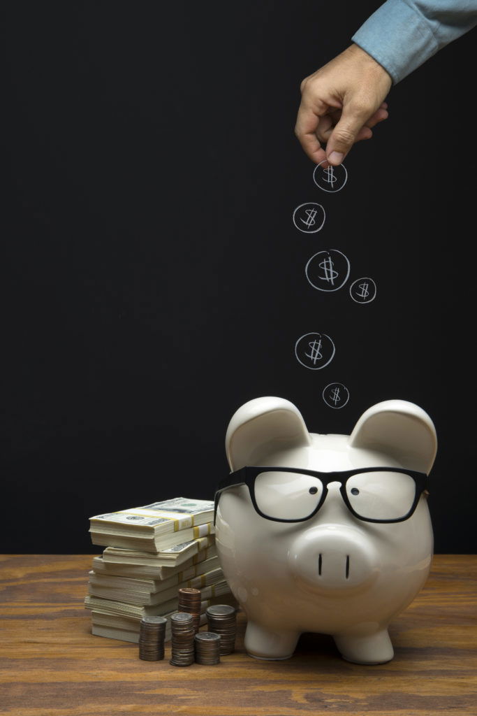 Large White Piggy Bank with hand depositing a chalk drawing money coin on Black Chalkboard Background This is a photograph of a large white Piggy Bank sitting on a desk a with a male hand depositing a chalk drawing of money coins on Black Chalkboard Background. This image could relate to savings and retirement.
