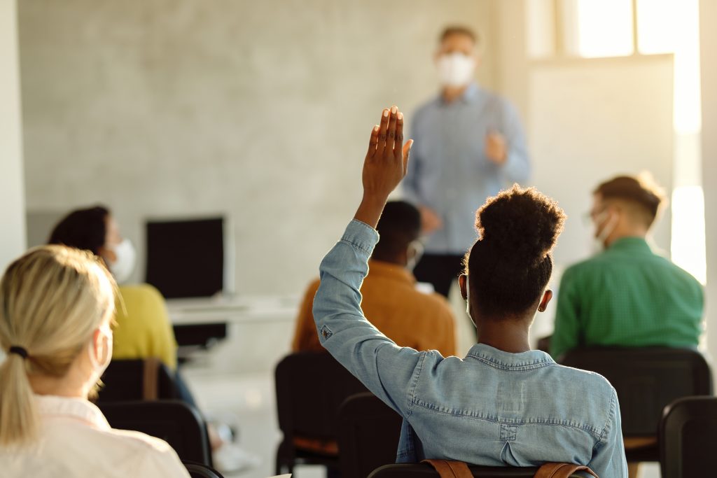 Back view of black female student raising arm to ask a question during a lecture in the classroom.