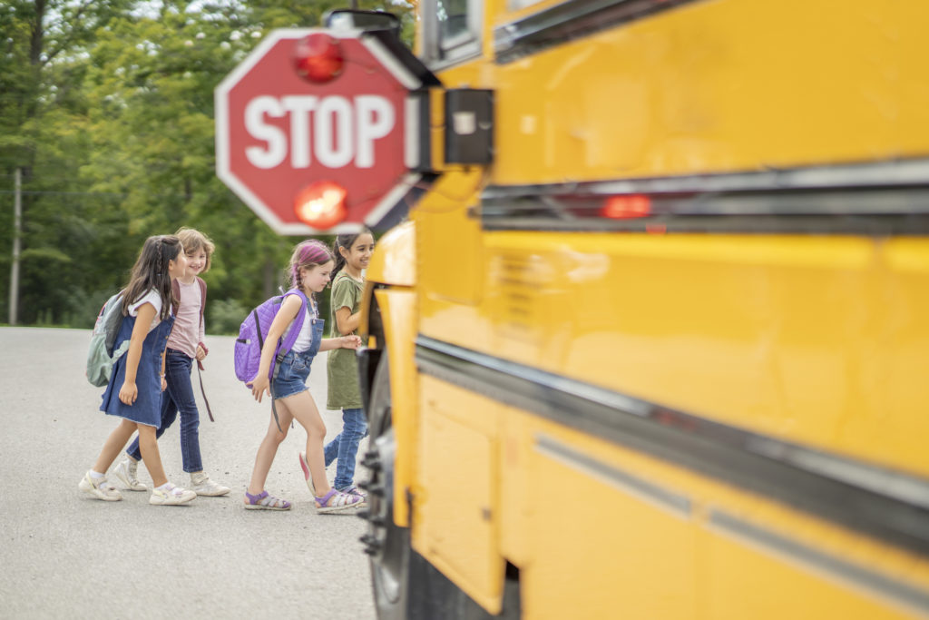 A yellow school bus is parked with its flashing Stop sign folded out as a small group of students cross in front.  The students are dressed casually and have backpacks on as they make their way to board the bus.